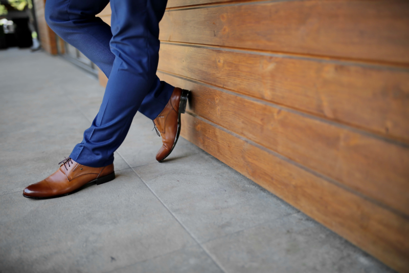 Homme en chaussures en cuir et pantalon de costume bleu marine, appuyé à un mur en bois